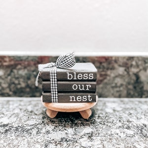 May include: Black wooden blocks stacked on a wooden stand with a black and white checkered ribbon. The blocks spell out "bless our nest".