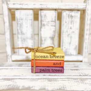May include: A stack of three small wooden blocks painted in shades of yellow, orange, and purple. The blocks are tied together with twine and have the text "ocean breeze and palm trees" written on them.