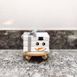 May include: A stack of white wooden blocks with a snowman face painted on them. The snowman has a black top hat, black eyes, and an orange carrot nose. The blocks are tied together with a black and white checkered ribbon.