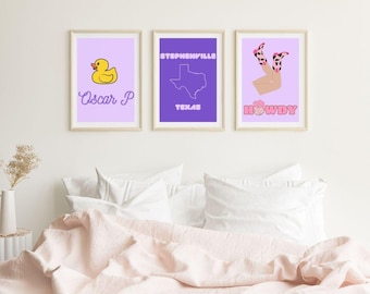 May include: Three framed prints on a white wall above a pink bedspread. The left print has a yellow rubber duck and the text "Oscar P". The middle print has a purple outline of the state of Texas and the text "Stephenville, Texas". The right print has a cartoon image of a woman's legs in pink and black boots and the text "Howdy".
