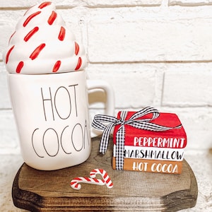 May include: White ceramic mug with the words "HOT COCOA" on the front, a red and white whipped cream lid, and a red and white candy cane on a wooden surface.  A stack of three wooden blocks with the words "Peppermint", "Marshmallow", and "Hot Cocoa" are next to the mug.