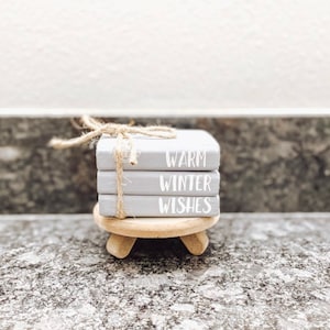 May include: A stack of three small gray books with the words "Warm Winter Wishes" written in white on the front. The books are tied together with twine and are sitting on a small wooden stand.