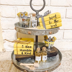 May include: A two-tiered metal tray with a rustic finish, decorated with honey-themed items. The top tier features a stack of yellow wooden blocks with the text "Sweet to the Soul", a small metal bucket with the word "Honey", a glass bottle with a honey bee label, and a wooden honey dipper. The bottom tier features a stack of yellow wooden blocks with the text "Bee Humble & Kind", two small glass bottles with gold glitter, and a metal bucket with the word "Honey".
