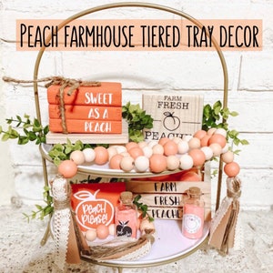 May include: A two-tiered gold metal tray with peach-themed farmhouse decor. The tray holds wooden blocks with text, peach-colored beads, small bottles, and decorative greenery. The text on the blocks reads "Sweet as a Peach" and "Farm Fresh Peaches".