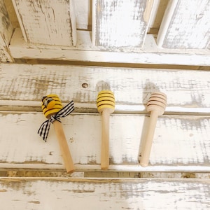 May include: Three wooden honey dippers with yellow and black beehive designs. The dippers are on a white wooden surface.