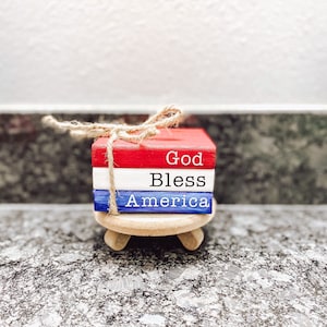 May include: A small wooden block set with the words "God Bless America" painted in red, white, and blue. The blocks are tied together with twine and sit on a small wooden stand.
