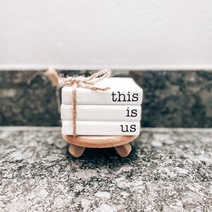 May include: White ceramic coasters stacked on a wooden stand with the words "this is us" written on them. The coasters are tied together with twine.