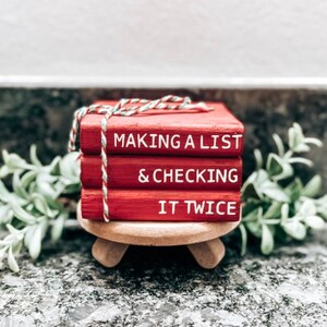 May include: Three red wooden books stacked on a wooden stand. The books are tied together with twine. The top book says "Making a List", the middle book says "& Checking", and the bottom book says "It Twice".
