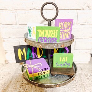 May include: A two-tiered metal stand with a handle, decorated with Mardi Gras themed wooden signs. The signs feature phrases like "Mardi Gras", "Beads and Bling", "Fat Tuesday", "King Cakes", and "Parades".