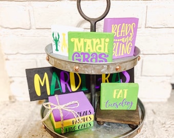 May include: A two-tiered metal stand with a handle, decorated with Mardi Gras themed wooden signs. The signs feature phrases like "Mardi Gras", "Beads and Bling", "Fat Tuesday", "King Cakes", and "Parades".