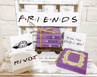 Op de afbeelding: Een set van zes Friends-thema houten onderzetters. De onderzetters bevatten het iconische Central Perk-logo, de zin "How You Doin?", de zin "Pivot", de zin "I'll be there for you" en een paars frame met een gele omtrek.