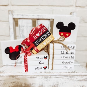 May include: A stack of white books with red and black polka dot ribbon. The books are decorated with Mickey Mouse and Minnie Mouse characters and the text "Home Sweet Home", "Mickey and Minnie", "Mickey", "Minnie", "Donald", "Goofy", and "Pluto".
