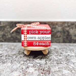 May include: A stack of three red wooden blocks with white lettering. The top block says "pick your own apples", the middle block says "5$ each", and the bottom block has a red and white checkered ribbon. The blocks are sitting on a small wooden stand with three legs.
