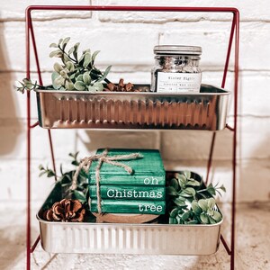 May include: A two-tiered metal shelf with a red frame. The top shelf has a jar of scented wax candle labeled "Winter Nights" and a small green plant. The bottom shelf has a stack of green books with the words "oh Christmas tree" and a small green plant.