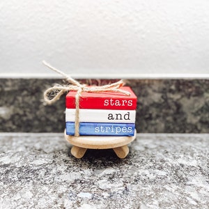 May include: A stack of three wooden blocks painted in red, white, and blue with the words "stars and stripes" written on them. The blocks are tied together with twine and are sitting on a small wooden stand.