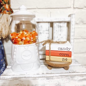 May include: A white gumball machine filled with candy corn sits next to a stack of three small books tied with twine. The books are painted white, orange, and yellow, with the words "candy corn 5¢" printed on them.