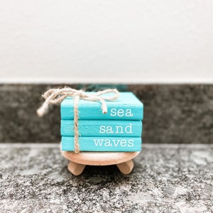 May include: Three stacked wooden blocks painted light blue with the words "sea", "sand", and "waves" written in white. The blocks are tied together with twine and are sitting on a small wooden stand.