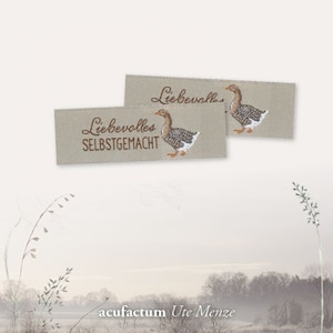 Puede incluir: Dos etiquetas de tela beige con bordado marrón de un ganso y el texto "Liebevolles Selbstgemacht". Las etiquetas están sobre un fondo blanco.