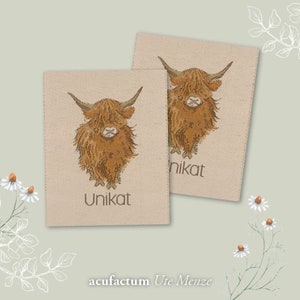Könnte beinhalten: Zwei braune Stoff-Etiketten mit einer Cartoon-Illustration einer Highland-Kuh. Die Etiketten sind beige mit braunem Text, der "Unikat" lautet.