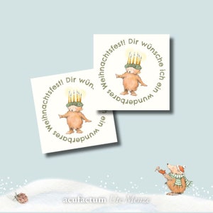 Pode incluir: Dois adesivos redondos com um urso marrom usando uma coroa de velas. O texto nos adesivos diz "ein wunderbares Weihnachtsfest! Dir wünsche ich", que se traduz como "Um Feliz Natal! Desejo-lhe". Os adesivos estão em um fundo azul claro com um pequeno urso marrom com uma trombeta em uma superfície nevada.