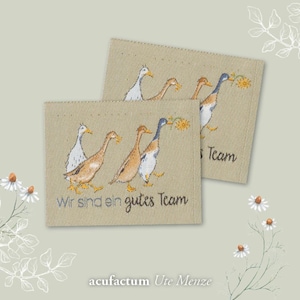 Könnte beinhalten: Drei Enten in einer Reihe, die letzte hält eine gelbe Blume. Die Enten sind braun und weiß mit blauen Köpfen. Der Text "Wir sind ein gutes Team" befindet sich unter den Enten. Der Text "acufractum Ute Menze" befindet sich unter dem Text.