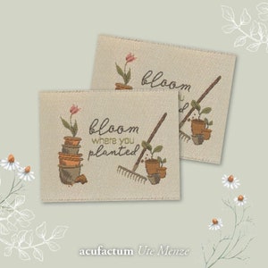 Pode incluir: Duas etiquetas de tecido bege com costuras marrons. A etiqueta superior diz "bloom" e apresenta uma flor rosa em um vaso. A etiqueta inferior diz "bloom where you planted" e apresenta uma flor rosa em uma pilha de vasos, um ancinho e uma planta em vaso. Ambas as etiquetas têm um design de ferramenta de jardinagem marrom.