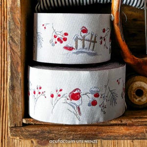 Puede incluir: Dos rollos de cinta de tela gris claro con un patrón tejido de pájaros de pecho rojo, bayas rojas y ramas nevadas. El texto Acufactum UTE MENZE está impreso en la parte inferior de la imagen en una fuente blanca sencilla sin remates.