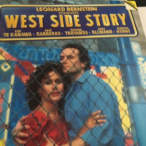 May include: A record album cover featuring a couple standing behind a chain-link fence. The album cover is for a recording of West Side Story conducted by Leonard Bernstein. The cast includes Kiri Te Kanawa, José Carreras, Tatiana Troyanos, Kurt Ollmann, and Marilyn Horne.