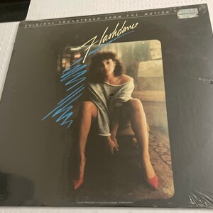 May include: Sealed vinyl record of the "Flashdance" soundtrack. The album cover features a woman in a gray sweatshirt and red shoes. The title "Flashdance" is in a stylized font with blue lines. The text "ORIGINAL SOUNDTRACK FROM THE MOTION PICTURE" is at the top.