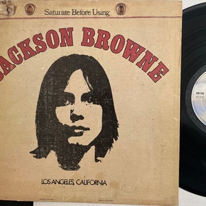 May include: A vintage vinyl record album cover featuring a black and white portrait of Jackson Browne. The album title "Jackson Browne" is in red and white lettering. The album cover also includes the text "Saturate Before Using" and "Los Angeles, California".