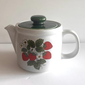May include: White ceramic teapot with a green lid and handle. The teapot features a strawberry design with red strawberries, green leaves, and white flowers. The teapot is a classic kitchenware item.