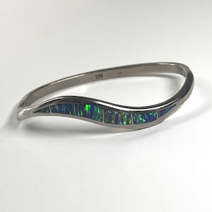 May include: A silver-toned hinged bangle bracelet featuring a wavy, curved design. The center of the band is inlaid with a series of rectangular, iridescent green and blue opal-like segments that create a vibrant, shimmering color pattern.