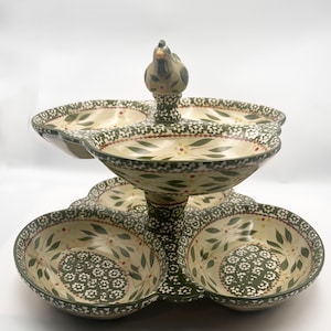 May include: A two-tiered ceramic serving dish featuring a cream-colored base with green leaf patterns, small white flower motifs, and red accents. The top tier has three sections and a central handle topped with a small sculpted bird figurine.