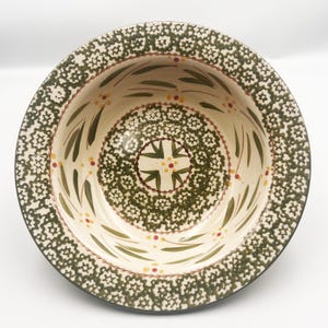 May include: A round ceramic bowl featuring a cream-colored base with a dark green floral pattern around the rim and center. The interior walls are decorated with green leaf motifs and small red and yellow dots arranged in a circular pattern.