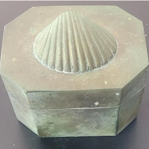 May include: An octagonal box with a weathered, muted green metallic finish. The lid features a raised, centered seashell motif with distinct radial ridges. The box has a uniform, aged patina across all visible surfaces, including the sides and top.