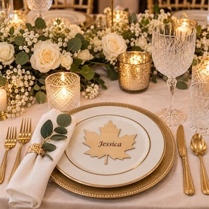 May include: A white dinner plate with a gold rim features a wooden maple leaf place card engraved with the name Jessica. The setting includes a white napkin with a gold ring, a sprig of eucalyptus, gold cutlery, and a crystal wine glass.