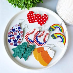 K&ouml;nnte beinhalten: Eine Sammlung von Ohrringen mit verschiedenen Designs, pr&auml;sentiert auf einem wei&szlig;en Teller. Die Designs umfassen rote Herzohrringe mit wei&szlig;en Punkten, Baseb&auml;lle, Regenbogen, Weihnachtsb&auml;ume und Zuckermais. Die Ohrringe sind aus leichtem Material gefertigt.