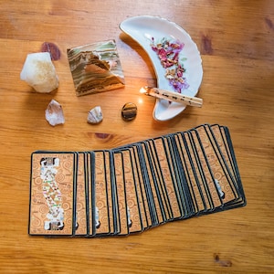May include: A deck of tarot cards with a black and gold design, a white crescent moon dish with dried flowers, a small piece of clear quartz, a small piece of brown and white agate, a small piece of citrine, a small piece of tiger's eye, and a wooden stick with a lit end.