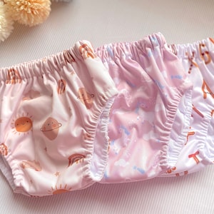 May include: Three pairs of pink and white reusable cloth nappies with different patterns. One pair has a sun and planets pattern, another has a unicorn pattern, and the last pair has a white background with a brown and white pattern.
