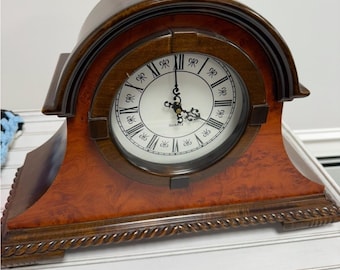 Puede incluir: Un reloj de cuarzo de madera con un diseño decorativo. El reloj tiene una esfera blanca con números romanos negros y manecillas ornamentadas. El marco de madera del reloj es una mezcla de tonos marrones oscuros y claros, con una parte superior curvada y una base detallada. El reloj mide aproximadamente 25 cm de ancho.