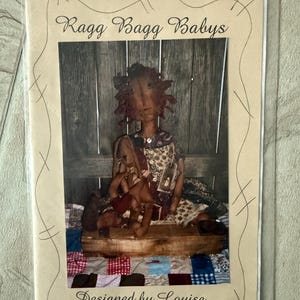 May include: A sewing pattern for a Ragg Bagg Baby doll, featuring a brown doll with a brown bunny sitting on a wooden bench. The pattern is designed by Louise.