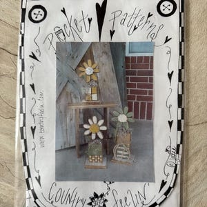 May include: A black and white illustration of a heart with the words "Pocket Patterns" inside. The illustration is surrounded by a black and white checkered border. The image also includes a photo of three wooden flower arrangements with a white daisy design. The photo is labeled "Country Feelin'" and includes a website address and phone number.
