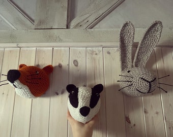 May include: Crocheted animal head wall decor. The image shows a fox, a badger, and a rabbit. The fox is orange and white, the badger is black and white, and the rabbit is gray. The animal heads are mounted on a light wood wall.
