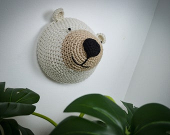 May include: A crocheted bear head wall hanging. The bear has a white body, a beige snout, and a black nose and mouth. The ears are small and rounded. The wall hanging is mounted on a white wall.