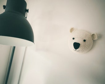 May include: A white, crocheted polar bear head fixed to a white wall. The bear has a black nose and eyes, with small, rounded ears. A black lamp is in the background.