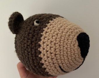 May include: A crocheted brown and beige bear head with black eyes and nose.
