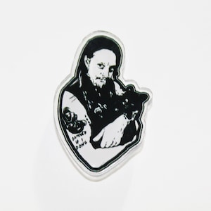 May include: Black and white sticker featuring a person with long hair and a beard holding a black cat. The person has a tattoo that says "DAMNED IF I DON'T". The sticker has a clear border.