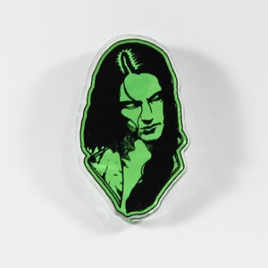 May include: Green and black acrylic pin featuring a portrait of a person with long black hair.