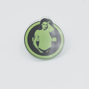 May include: A green and black round button with a graphic of a man with long hair. The man is wearing a black shirt and has a green background.