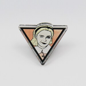 May include: A triangular acrylic pin with a black border. The pin features a cartoon image of a woman with blonde hair, wearing a white collared shirt and winking. The background is a peach color.